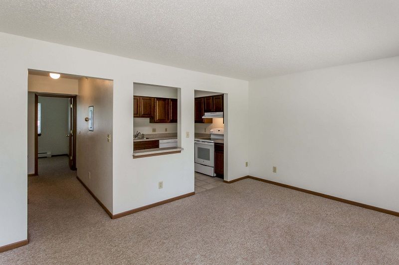 Empty beige carpeted living room with pass-through to a kitchen and hallway