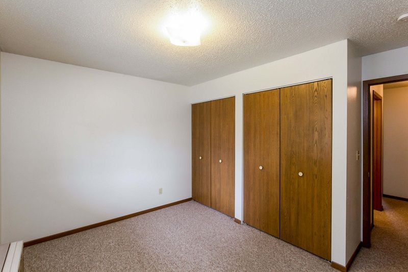 Empty beige bedroom with white walls, carpet, and wooden closet doors near a hallway entrance