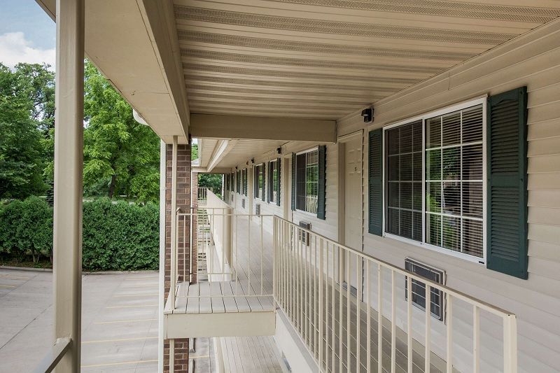 Covered apartment walkway with white railings, beige walls, and green shutters beside a parking area.