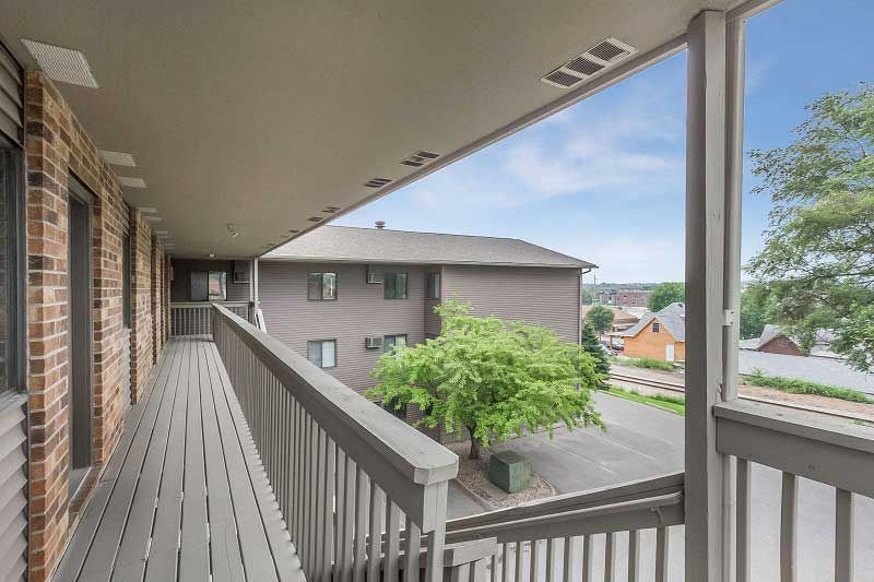 Covered apartment balcony with gray railings overlooking neighboring buildings and trees