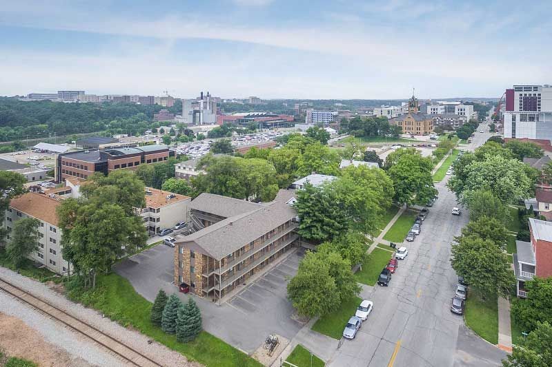 Aerial view of a city street with trees, buildings, and parked cars under a cloudy sky
