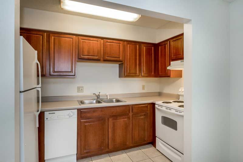 Compact kitchen with wooden cabinets, white appliances, and beige tile flooring