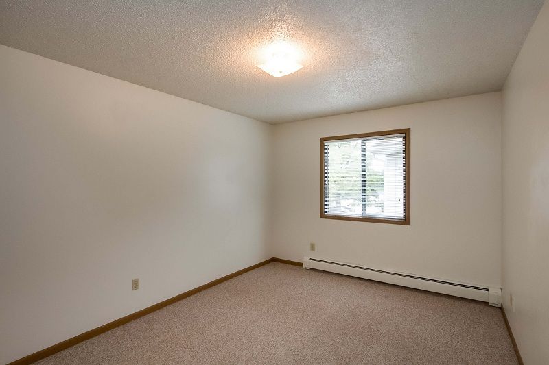 Empty beige bedroom with carpet, window, and baseboard heater.