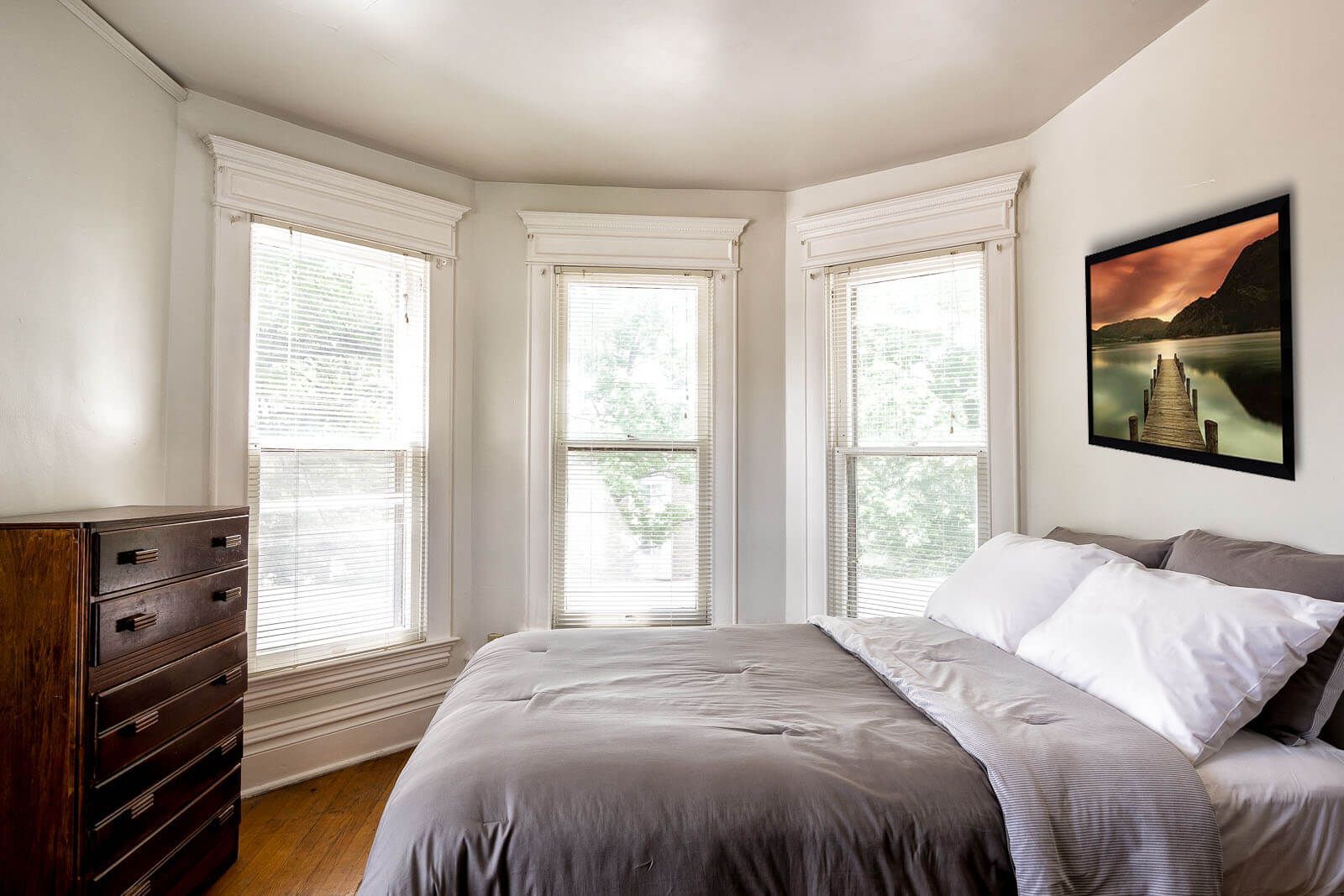 Bright bedroom with three bay windows, gray bedding, a dresser, and a landscape painting.