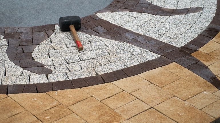 A mallet rests on a patterned brick walkway under construction; brown, beige, and white bricks are visible.