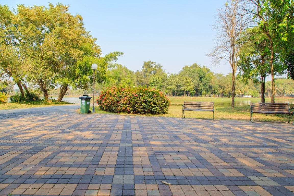 Park scene with brick walkway, trees, a bench, and trash can.