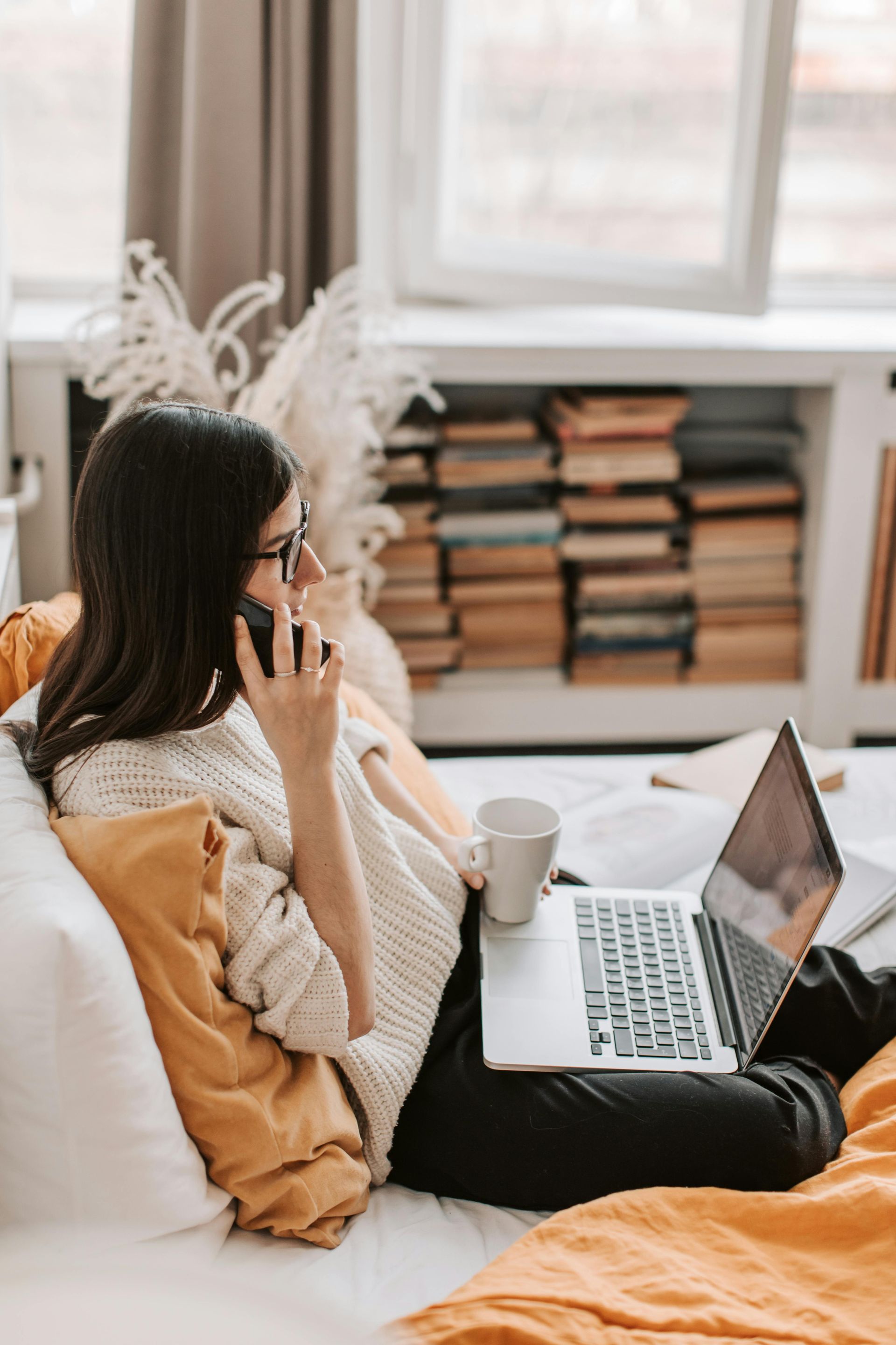 A woman is sitting on a bed with a laptop and talking on a cell phone.