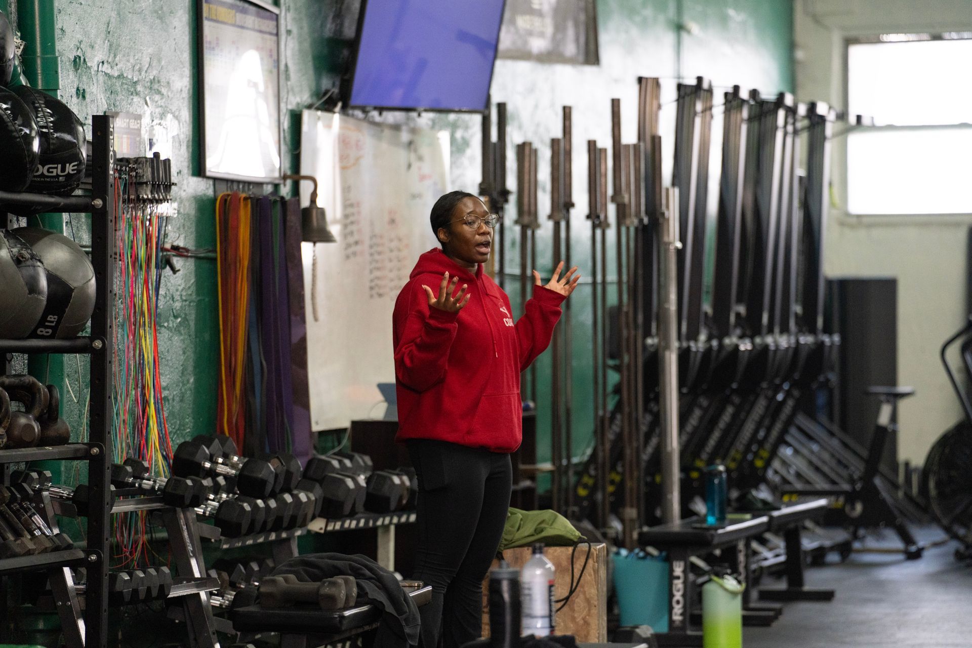 Person in a red jacket gesturing in a gym with weight racks and equipment along the wall