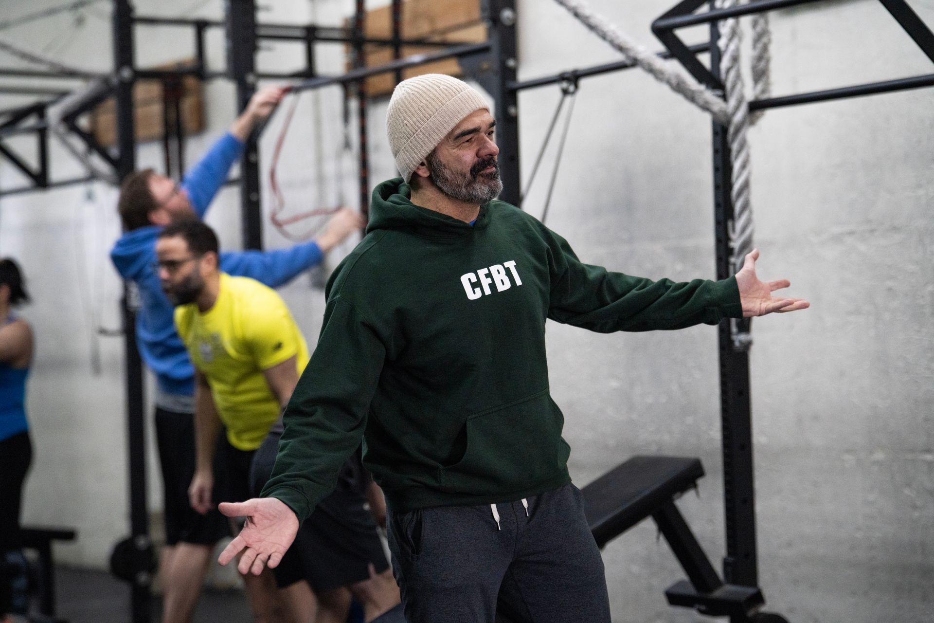 Man in green hoodie and knit beanie gesturing in a gym with workout rigs, others blurred in background
