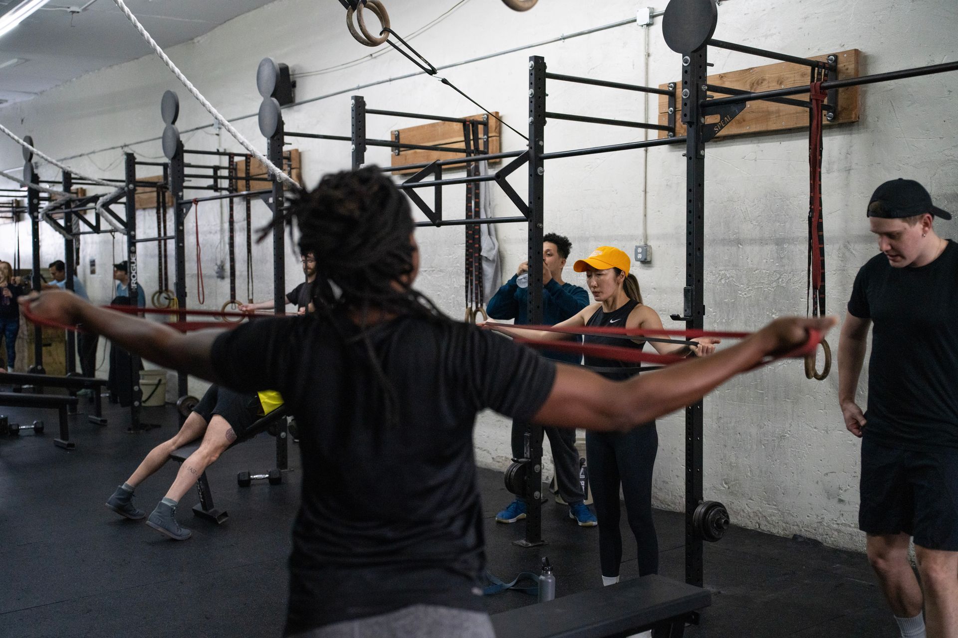 People exercising in a gym with pull-up rigs; one person stretches arms wide in the foreground.