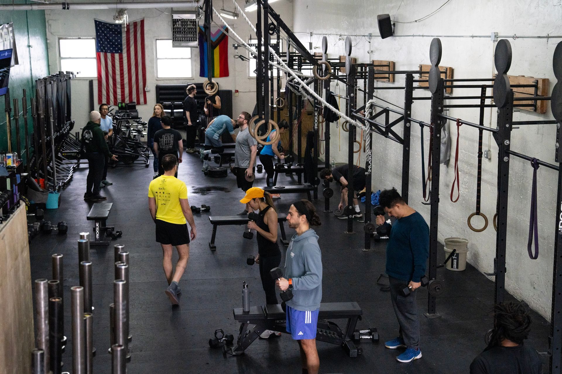 People exercising in a gym with dumbbells, pull-up rigs, and flags on the wall.