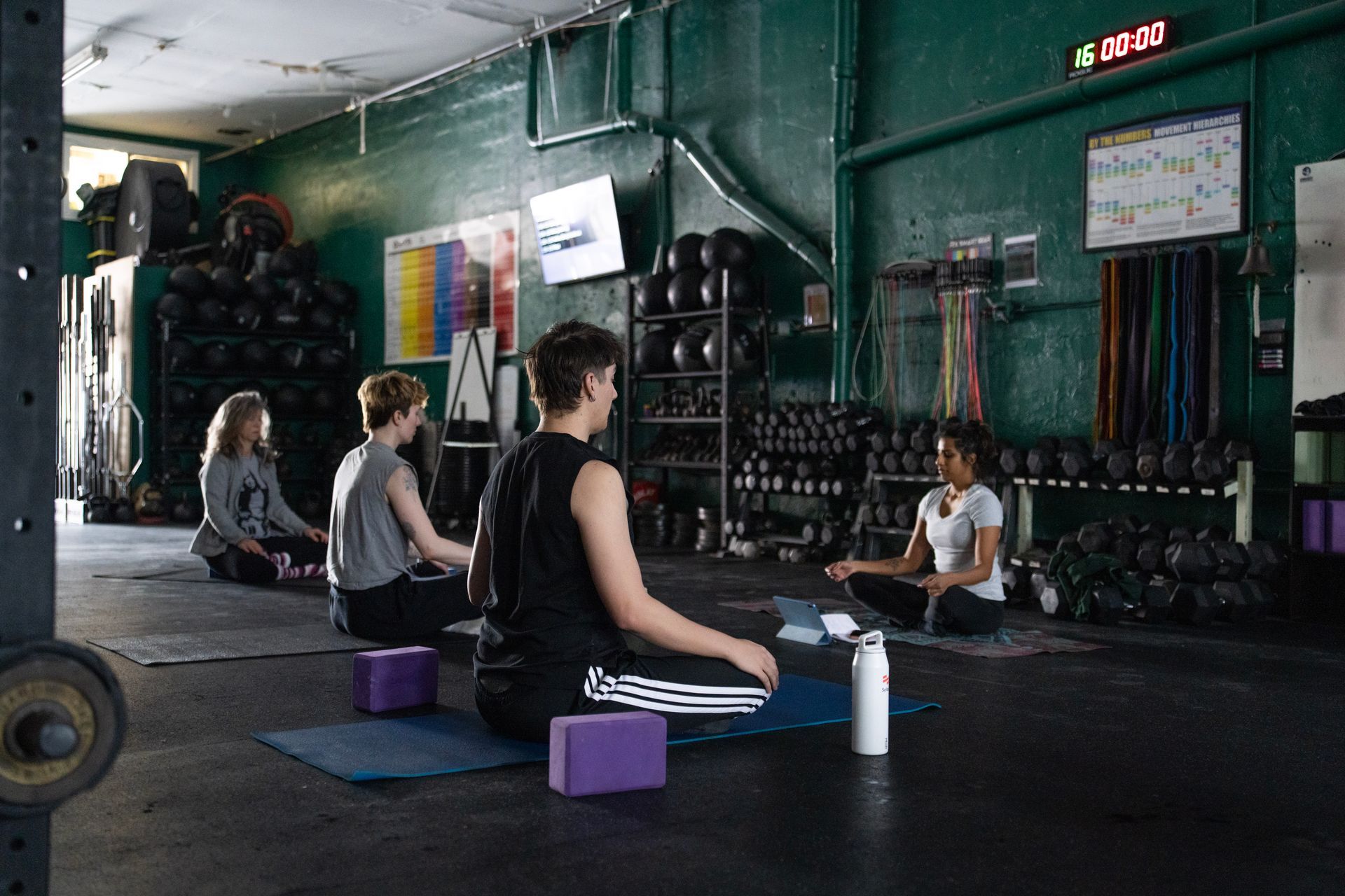 People doing yoga stretches in a gym with mats, blocks, dumbbells, and wall-mounted equipment