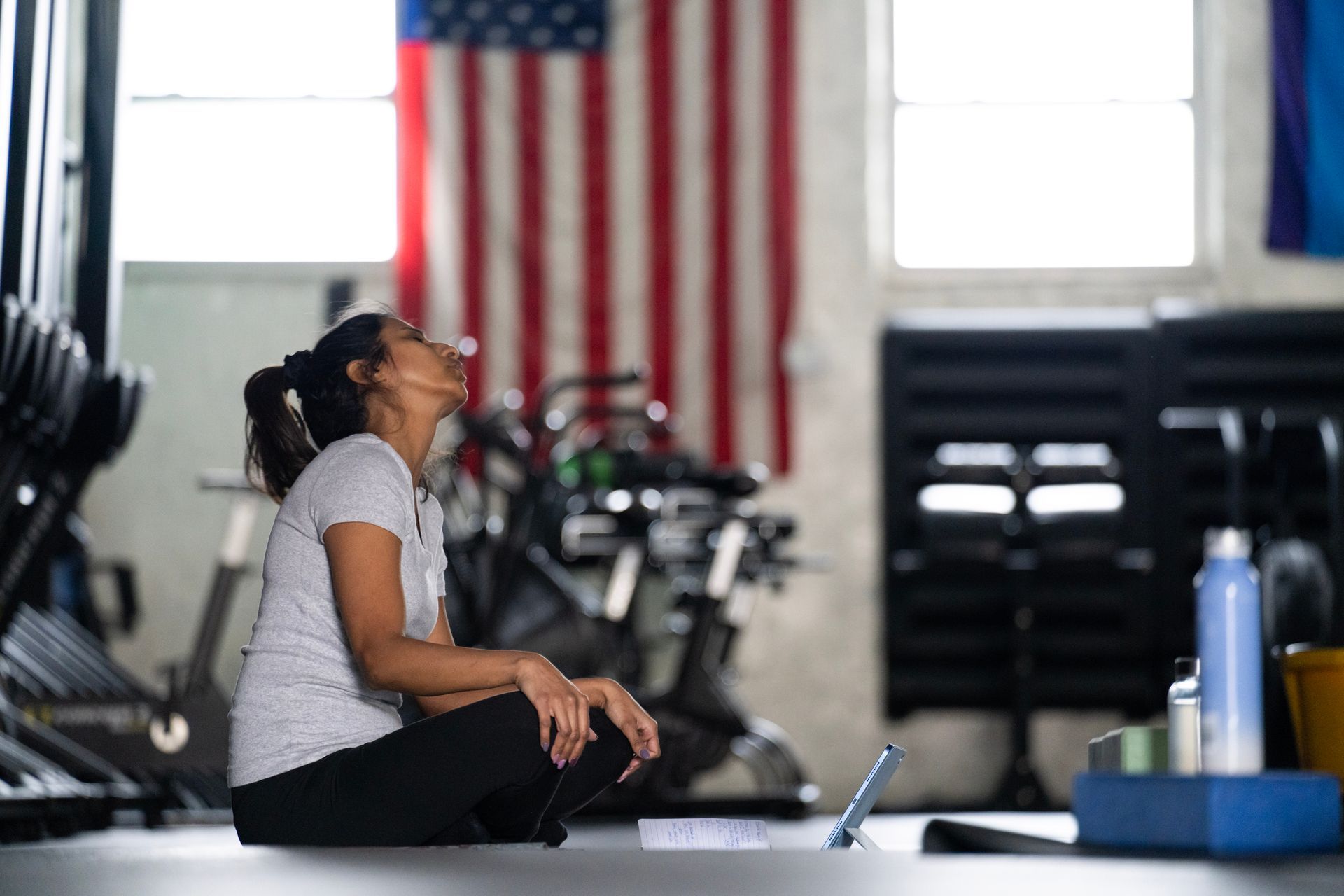 Person resting on a gym floor with workout equipment and an American flag in the background