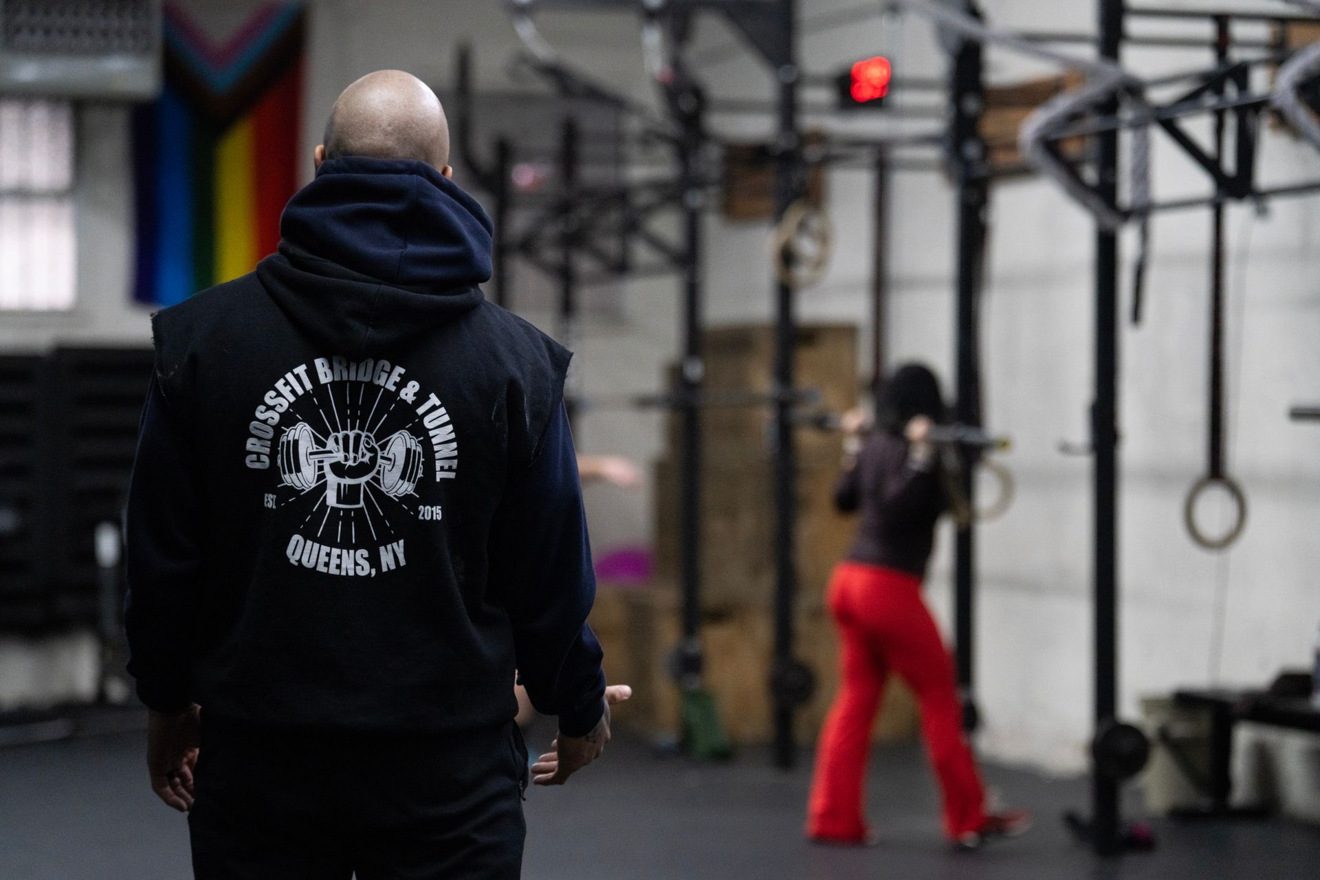 Person in a black hoodie faces a gym where another person in red pants practices near gym rings.