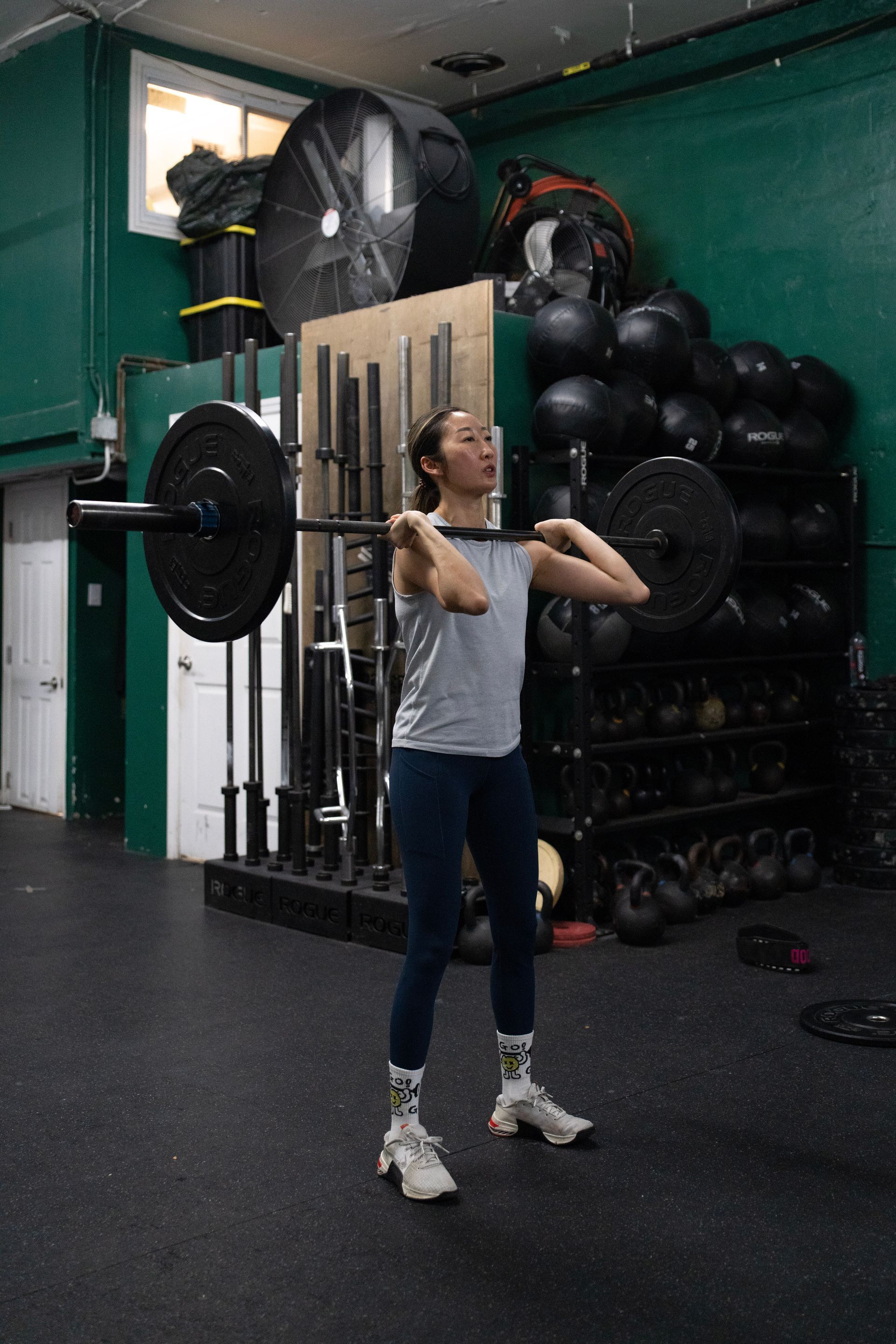 Woman holding a barbell in a gym, with medicine balls and weights stacked behind her.