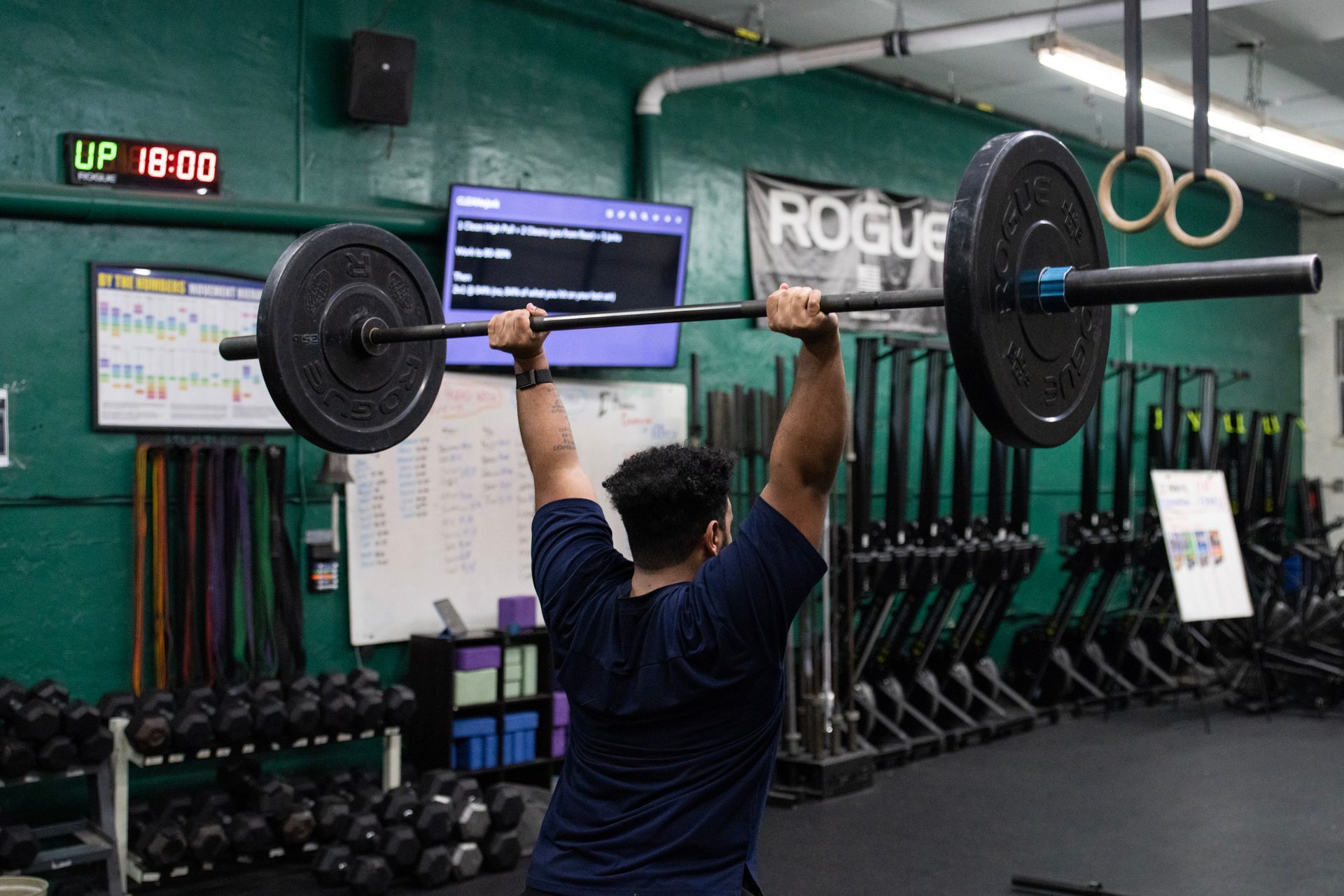 Person lifting a barbell overhead in a gym with a mirror, screens, and a digital timer in the background