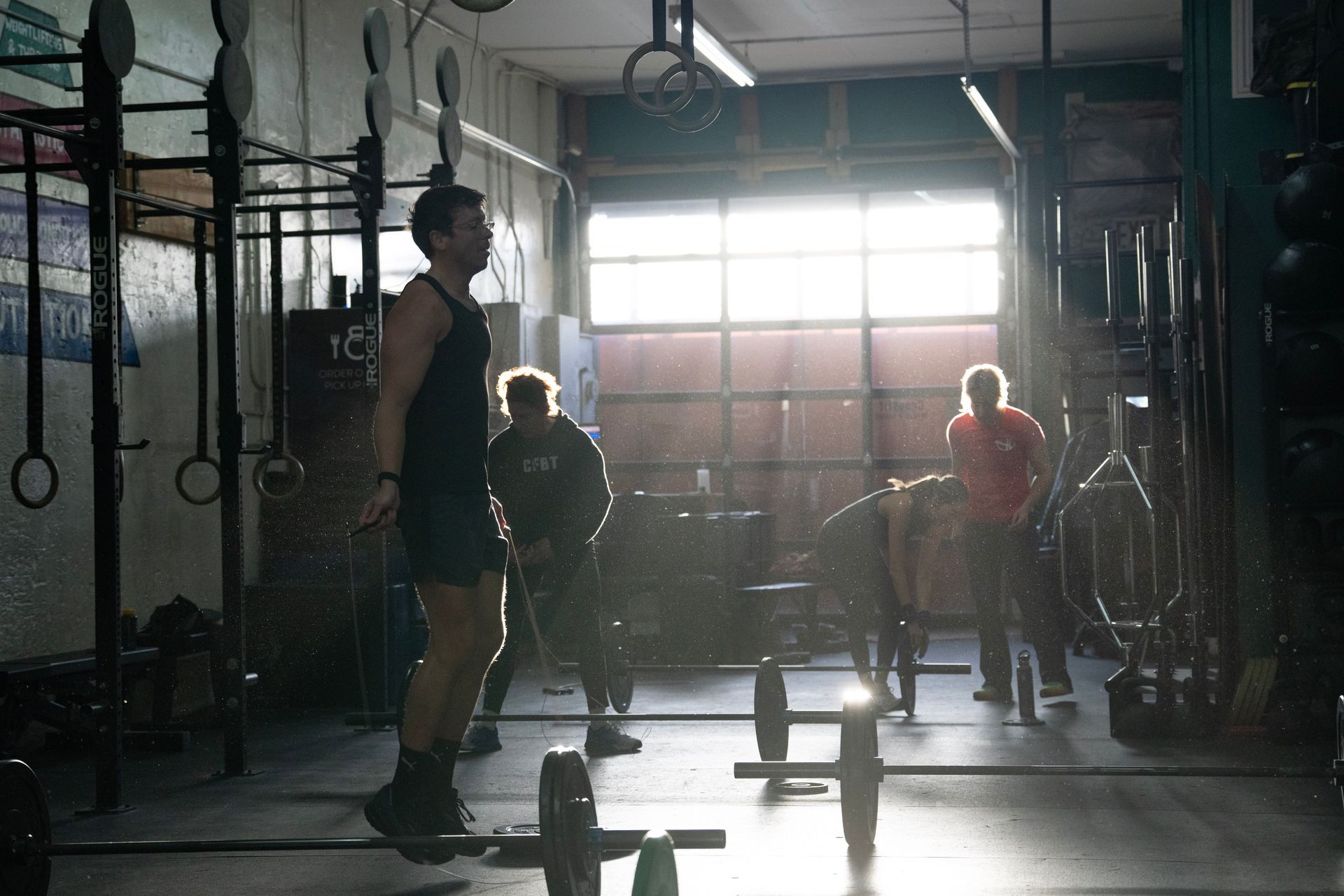 People in a gym with barbells and rings, lit by bright daylight through large windows.