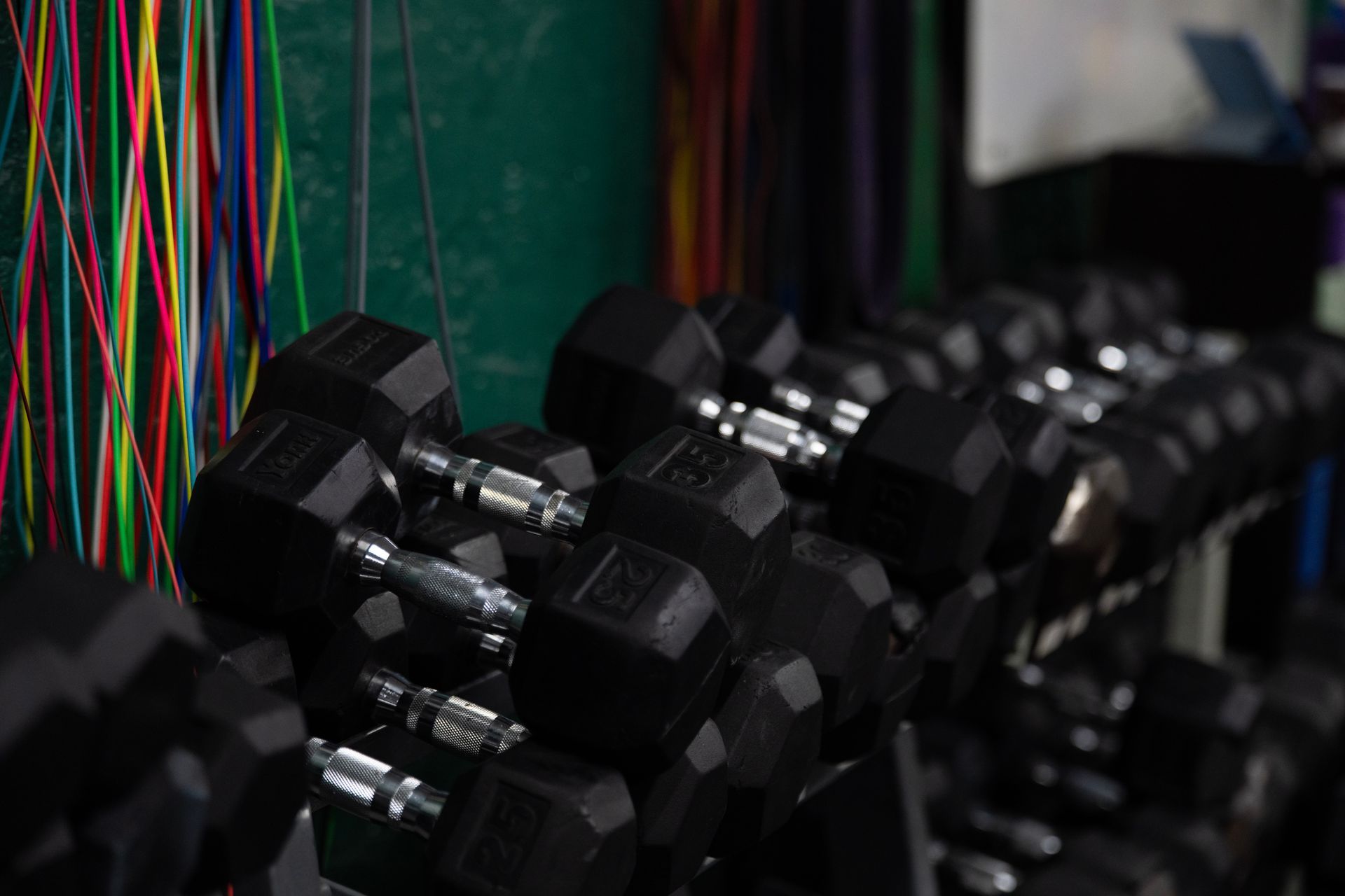 Row of black hex dumbbells with colorful resistance bands hanging behind them in a gym setting