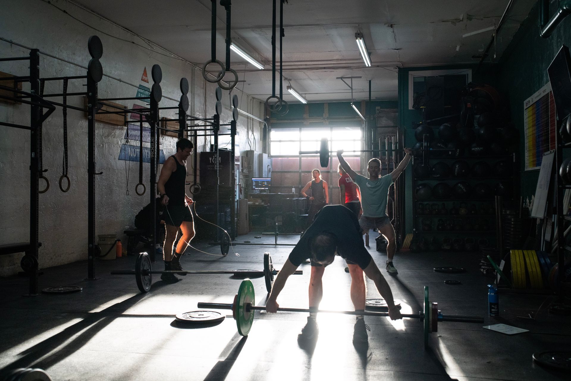 People doing barbell workouts in a dim CrossFit gym with sunlight streaming in.