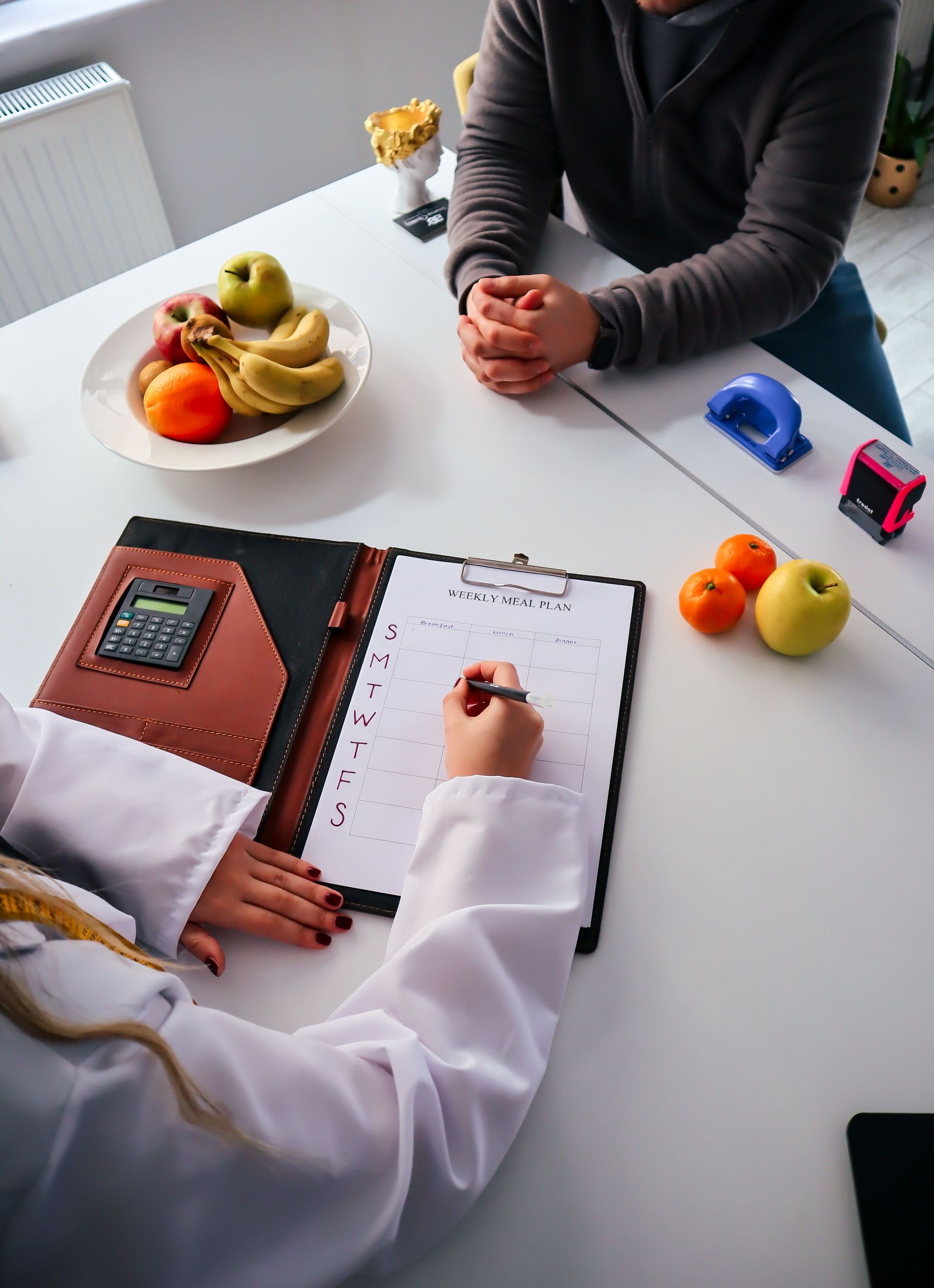 Two people sit across a desk with an open notebook, fruit bowls, and small office items.