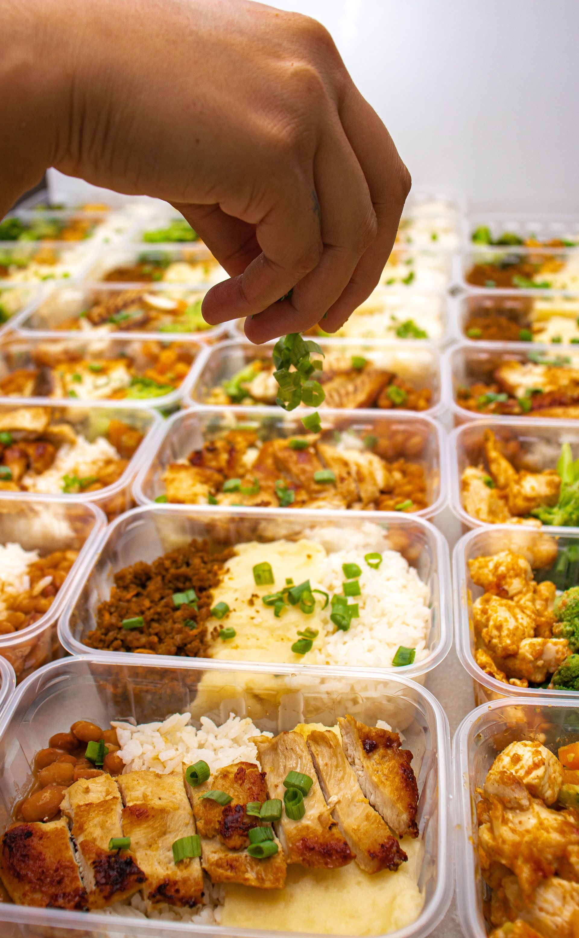 Hand placing garnish on rows of meal-prep containers with rice, vegetables, and chicken