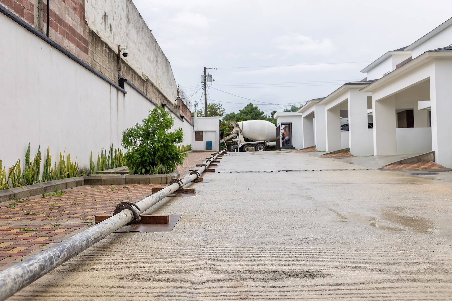 Concrete driveway beside white buildings, with a large pipe along the left side and a cement mixer in the distance.