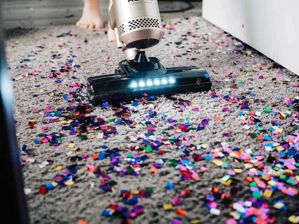 Vacuum cleaner head sucking up colorful confetti from a carpet beside bare feet