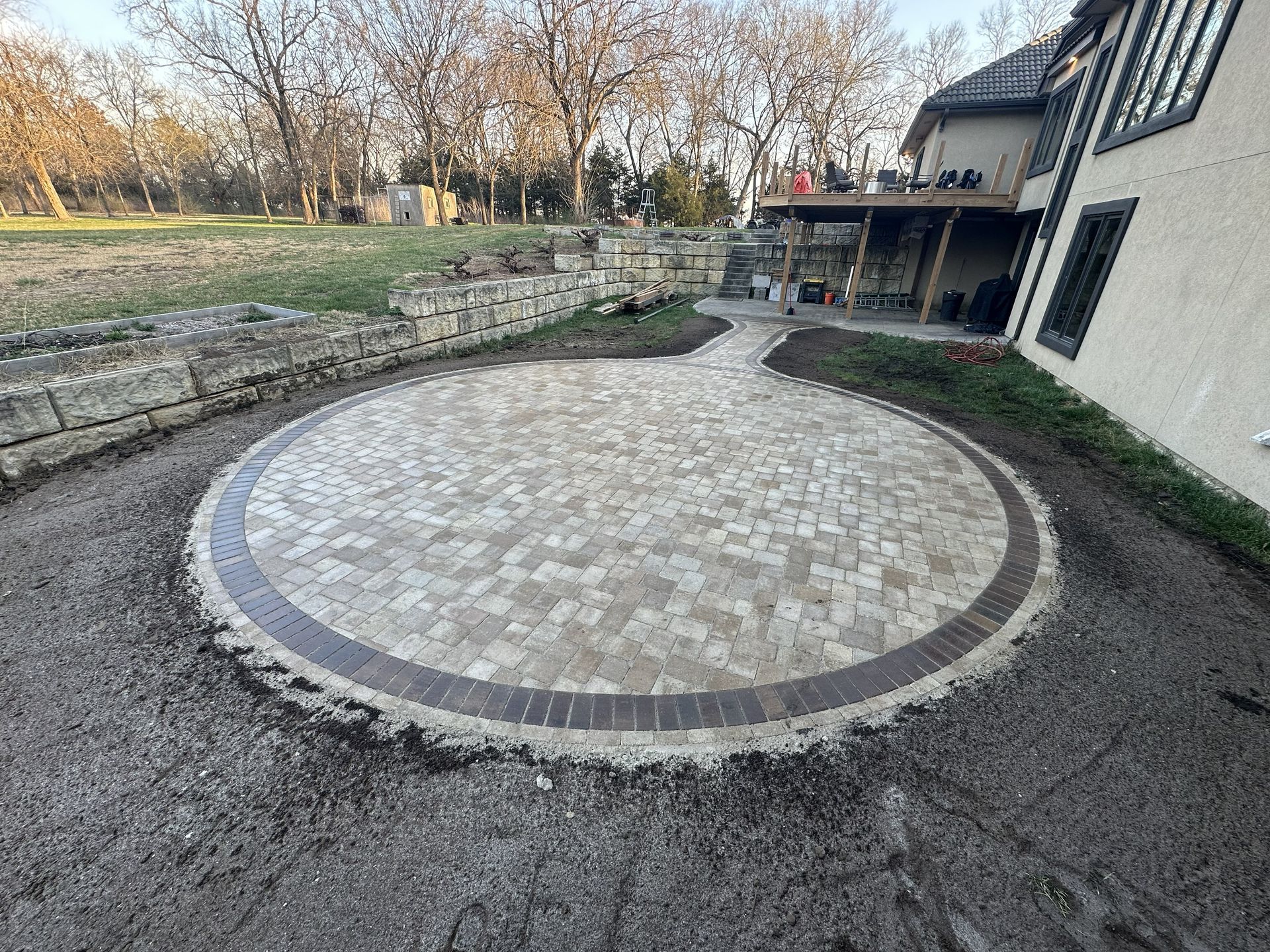 Circular stone patio beside a house, bordered by a low stone wall and surrounded by trees.