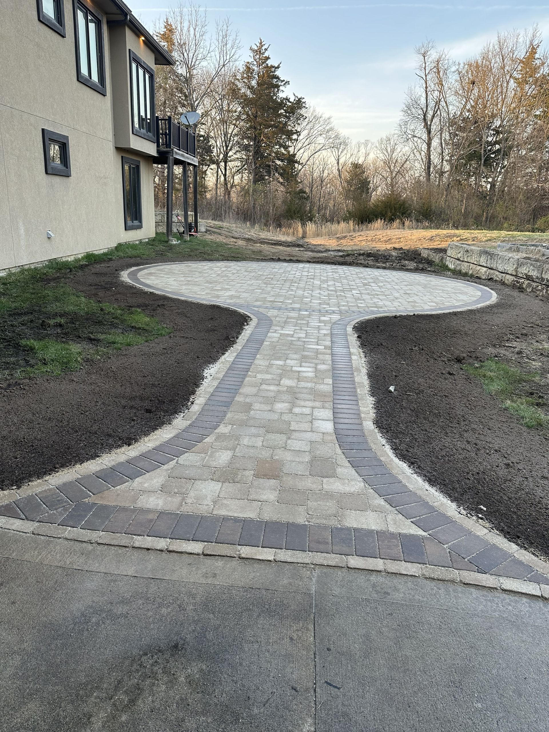 Curved stone walkway beside a house, leading through a landscaped yard to trees.
