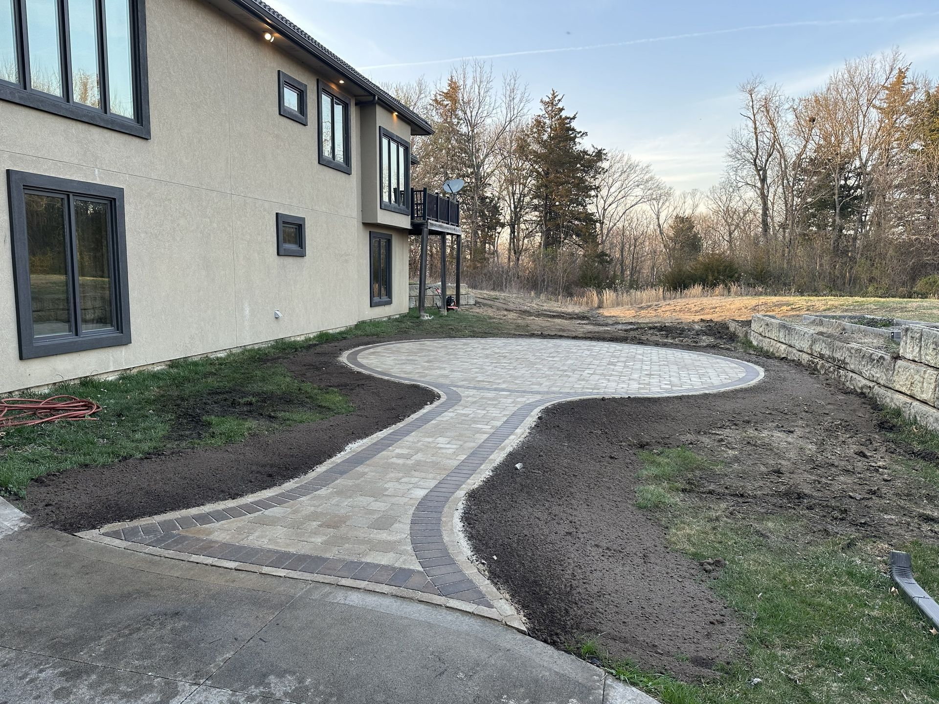 Curved stone pathway beside a house, bordered by soil and a low stone wall in a garden yard.