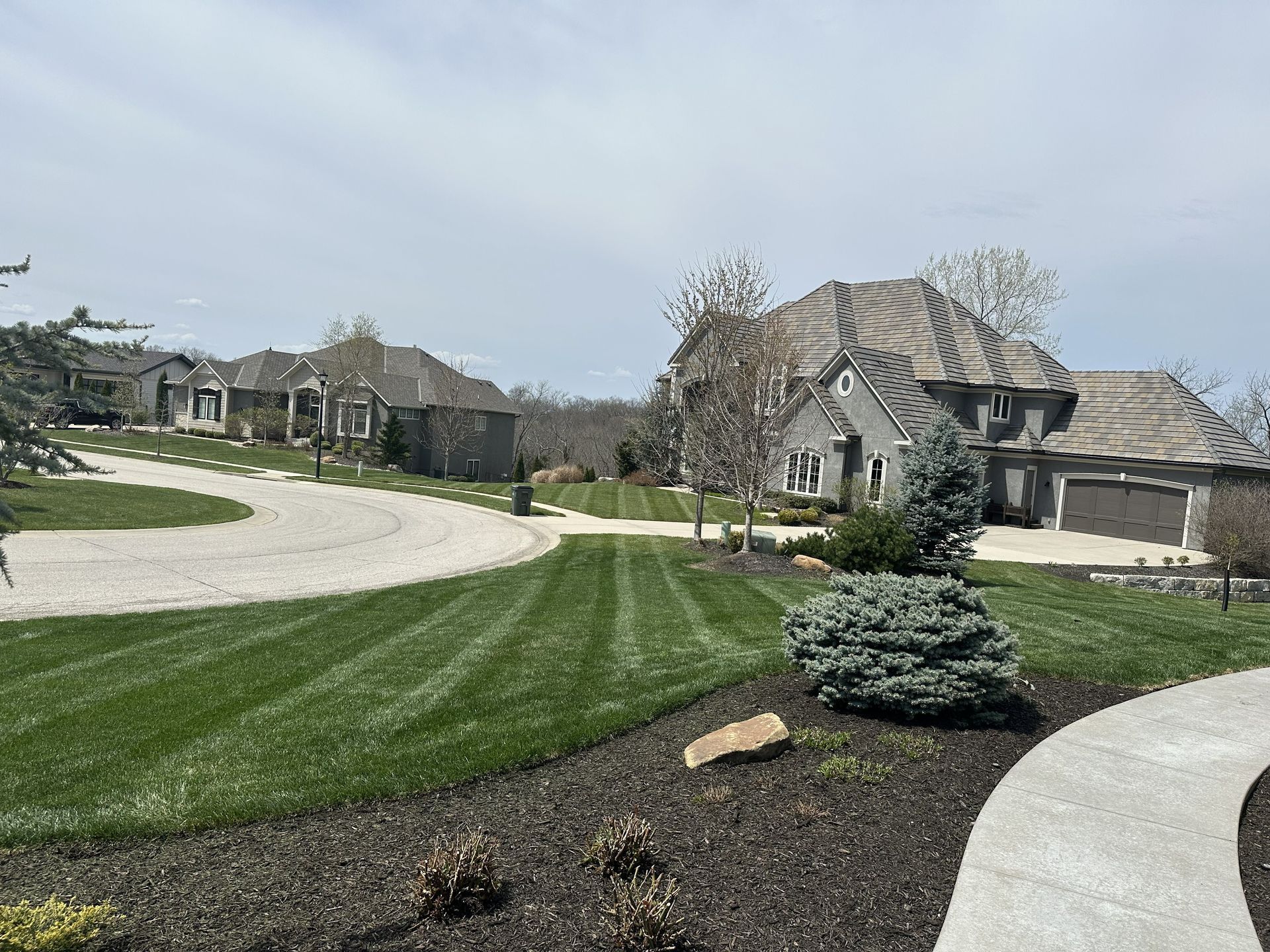 Suburban neighborhood with large houses, curving sidewalks, and freshly landscaped lawn under a cloudy sky
