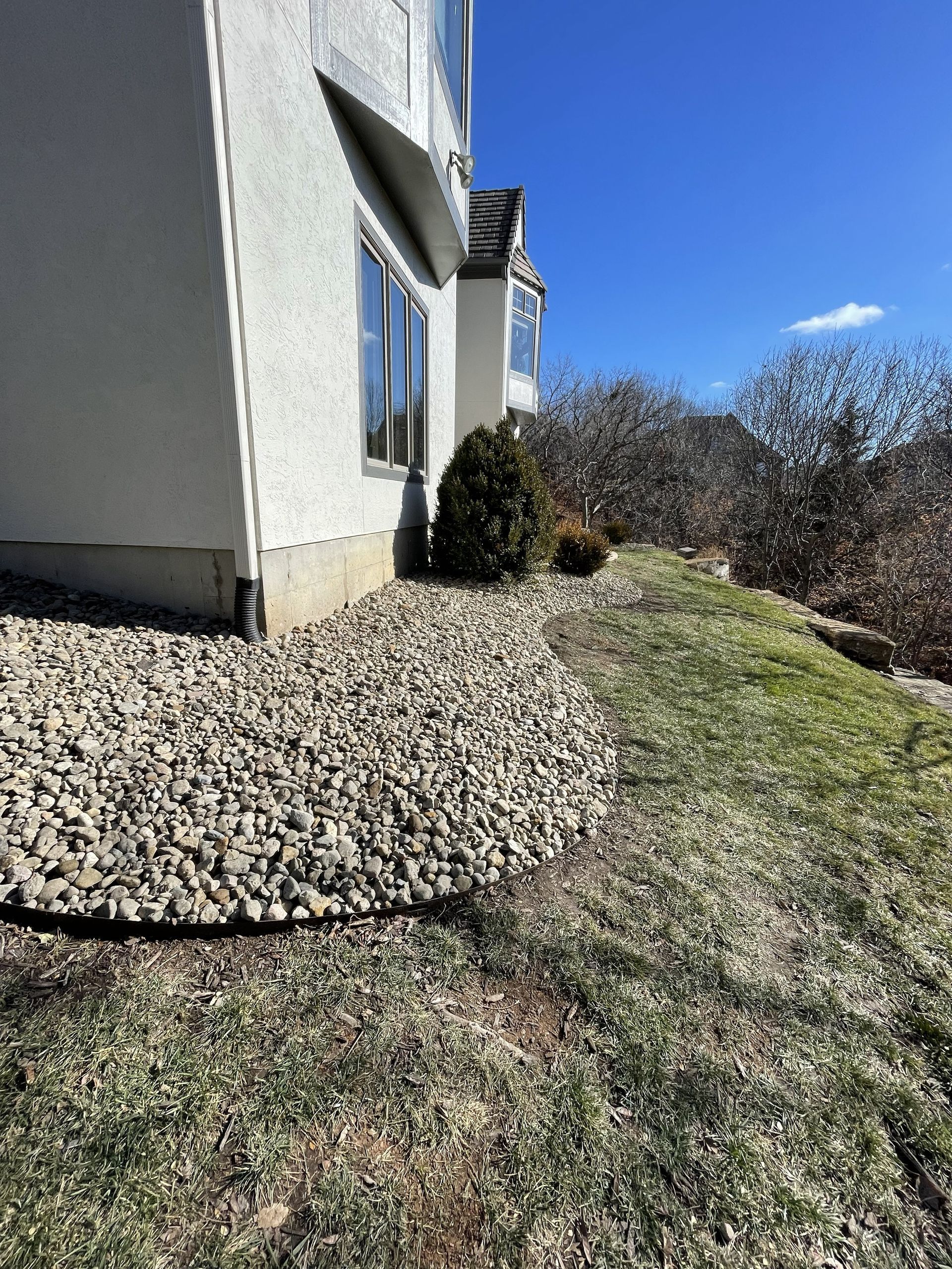 Side of a house with a stone border, grass lawn, shrubs, and blue sky on a sunny day