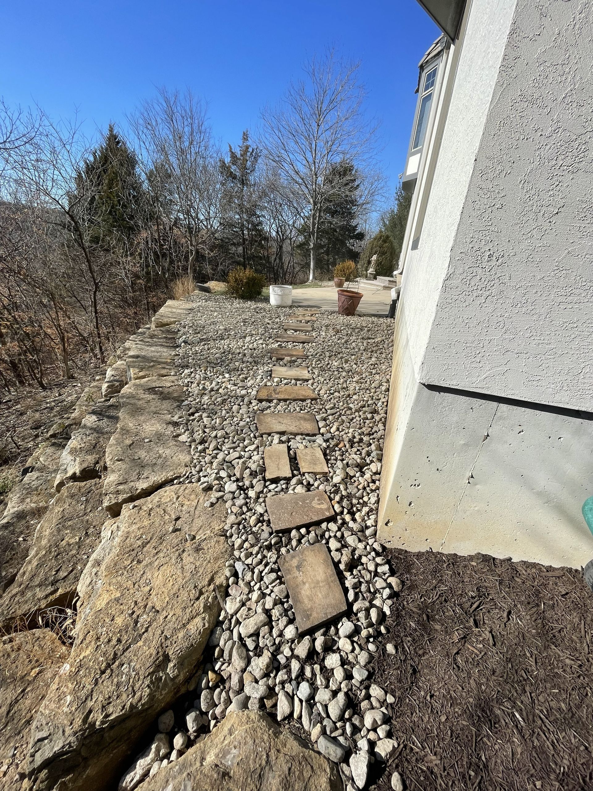 Gravel path with stepping stones beside a white building, bordered by large rocks and trees under a blue sky
