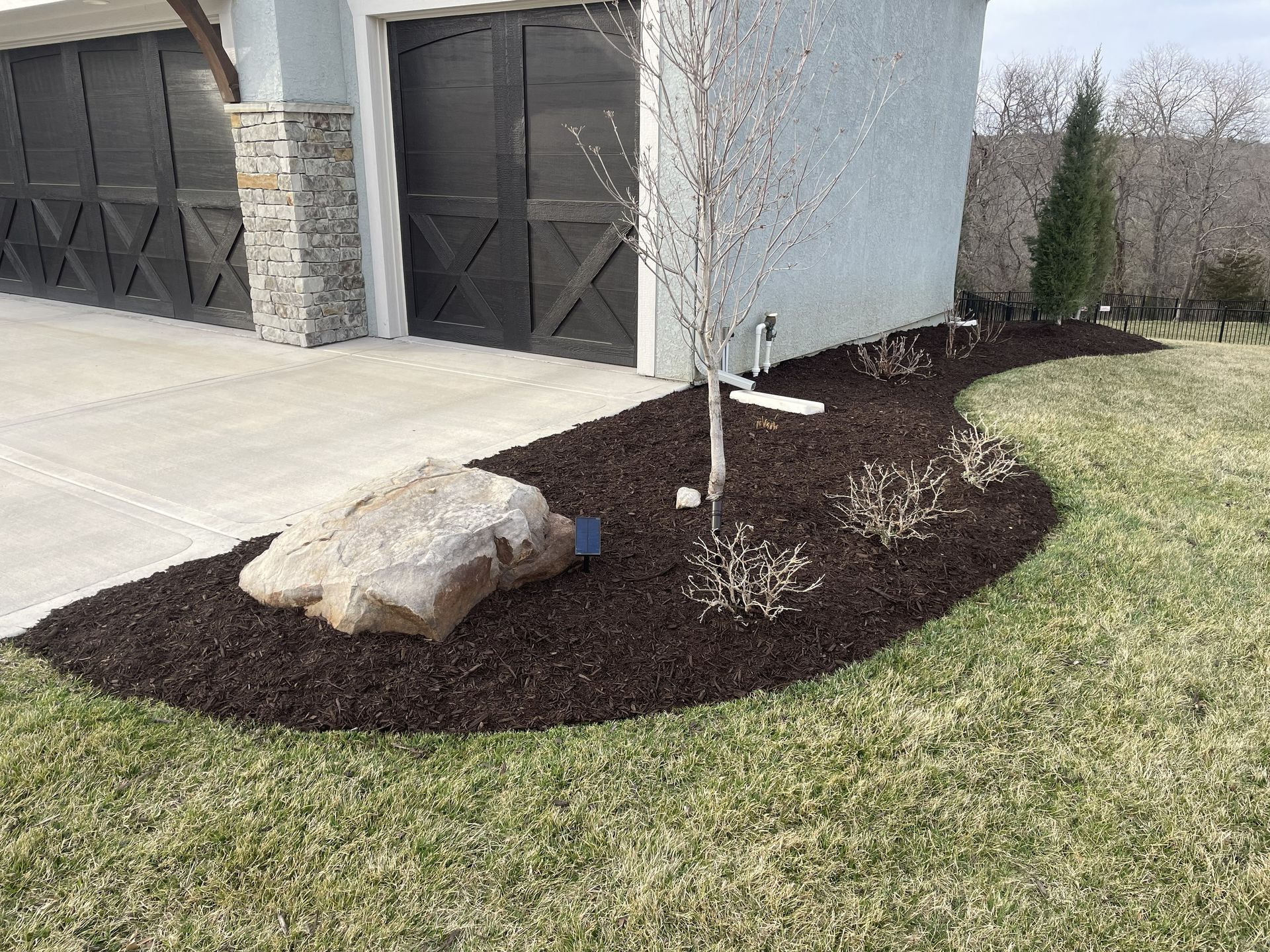Landscaped front yard with mulch, large rocks, a young tree, and garage doors beside a driveway