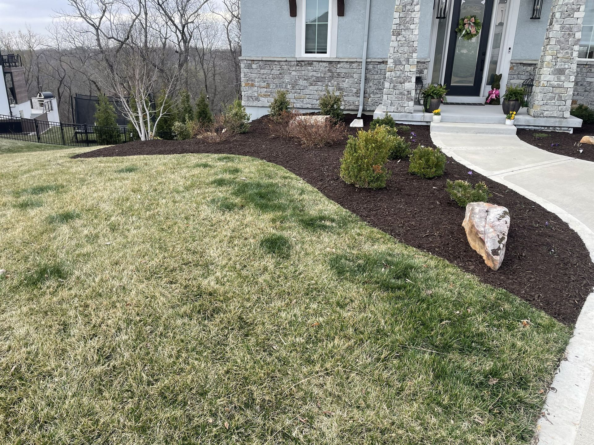 Front yard with patchy grass, mulched curved garden bed, small shrubs, and stone front walkway by a house