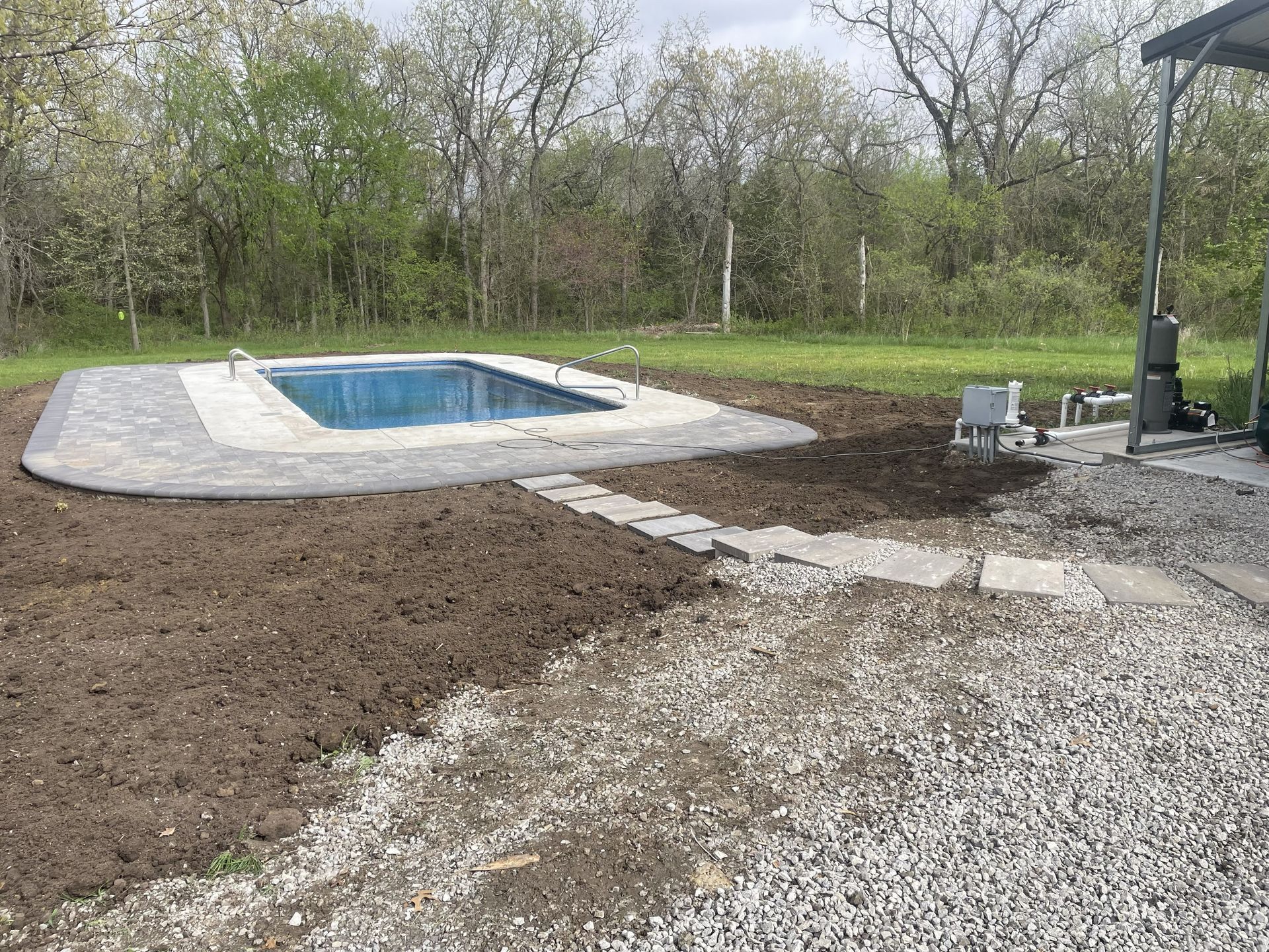 Backyard pool under construction with gravel, dirt, and a stone walkway beside trees.