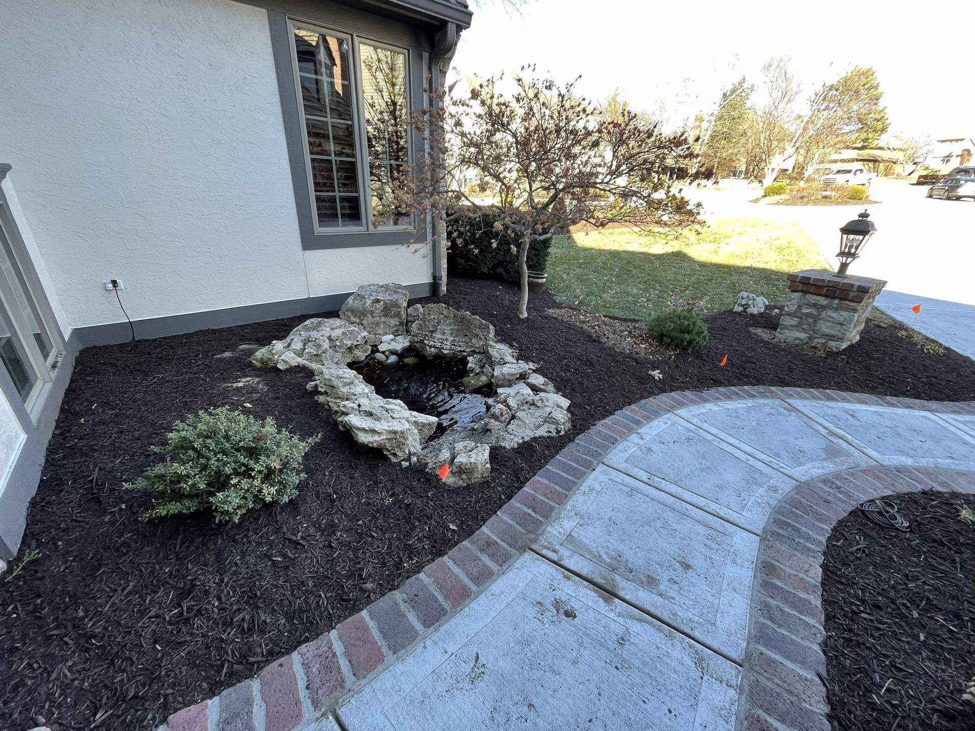 Curved brick walkway beside a house, landscaped with rocks and shrubs, leading to a yard and driveway.