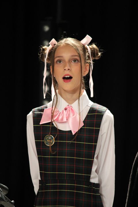 Girl with hair buns, ribbons, school uniform, singing, black background.
