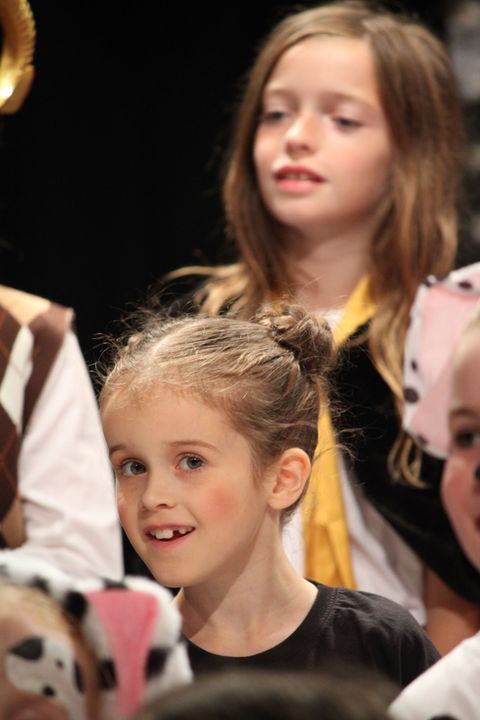 Children in costumes, singing on stage. Front girl smiles, other has a neutral expression.