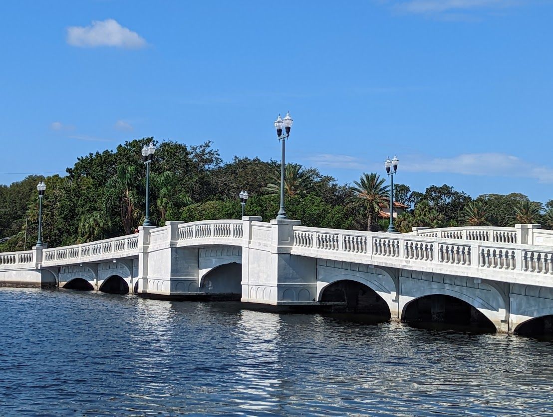 White arched bridge over blue water under a clear sky, with lampposts and trees in the background