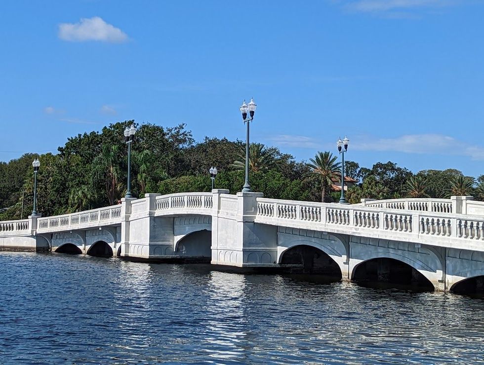 White arched bridge over blue water under a clear sky, with lampposts and trees in the background