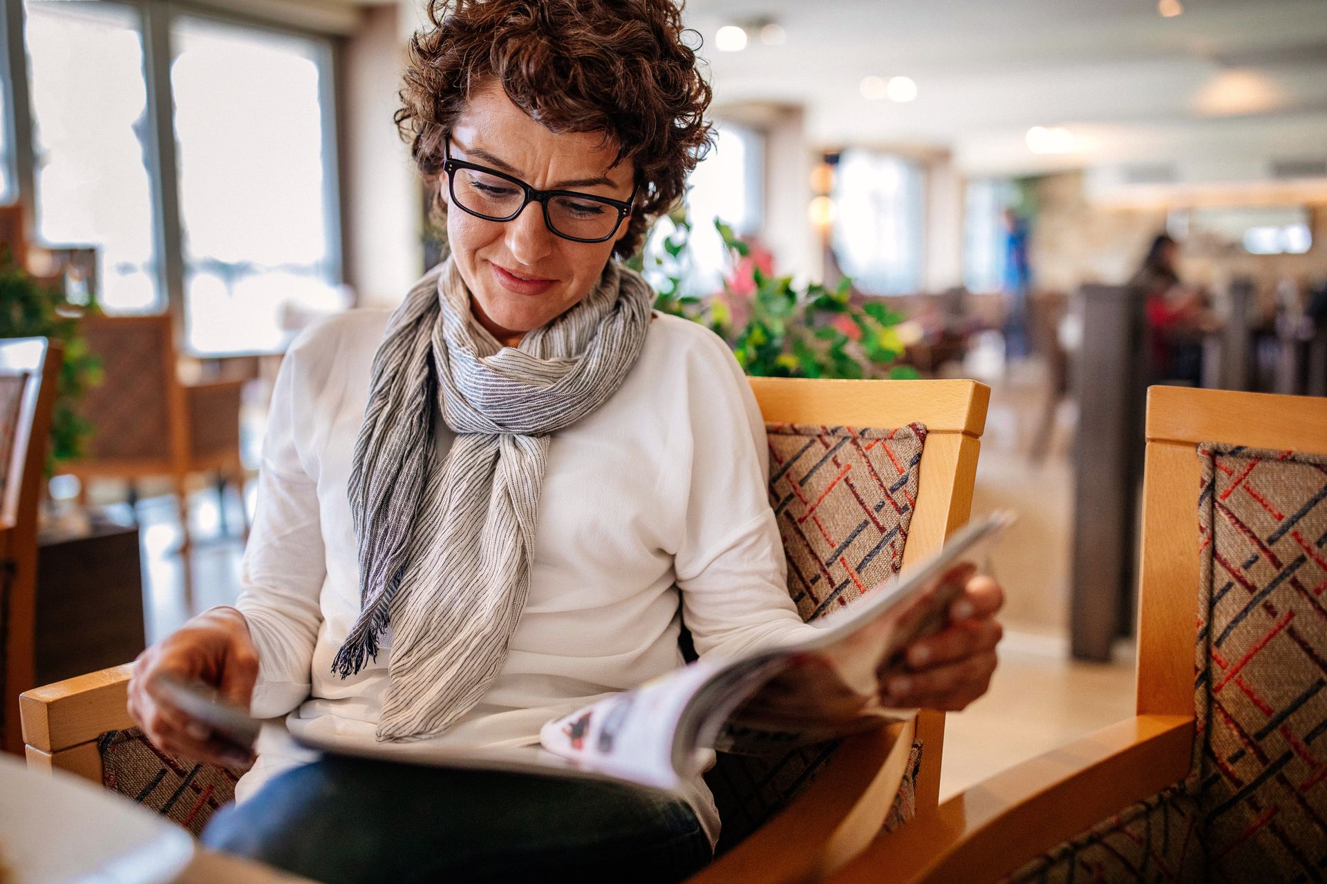 Person reading a magazine in a cozy cafe, seated by a window with plants and patterned chairs.