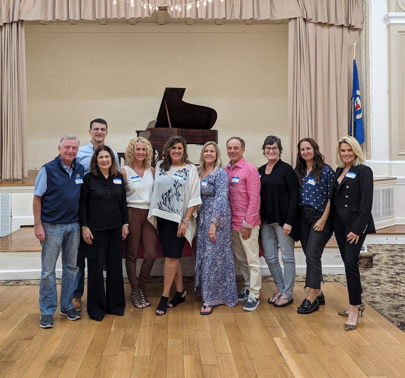 Group posing on a stage in front of a grand piano, with curtains and a blue flag in the background.