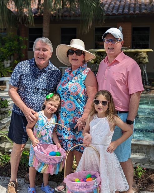 Four people and two girls pose outdoors by steps, holding pink Easter baskets.