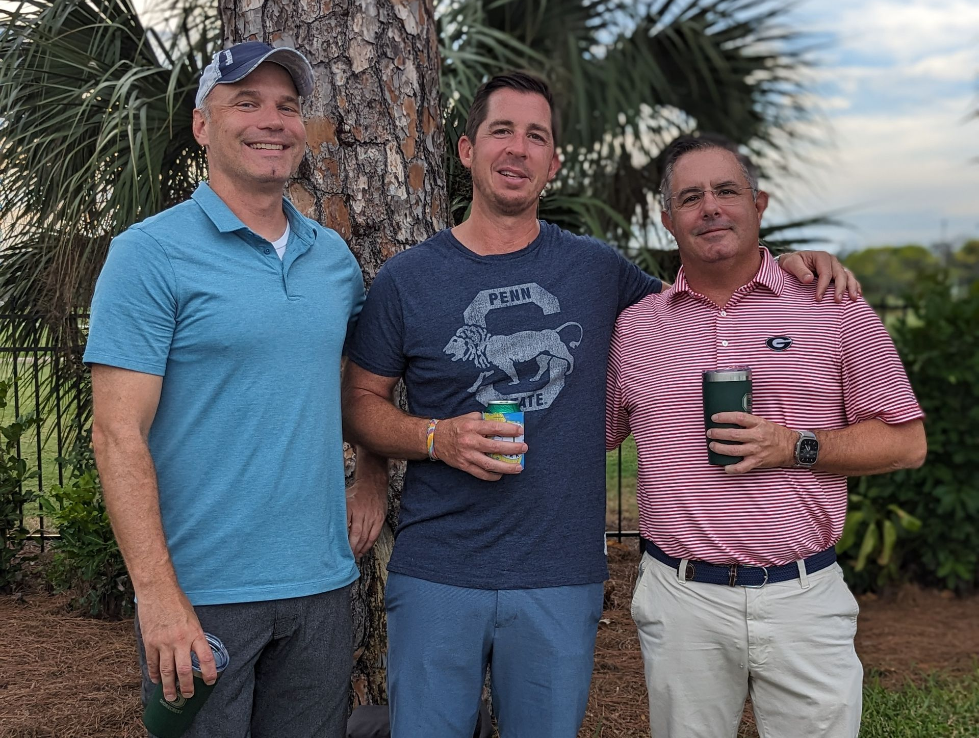 Three men standing outdoors by a palm tree, smiling and holding drinks.