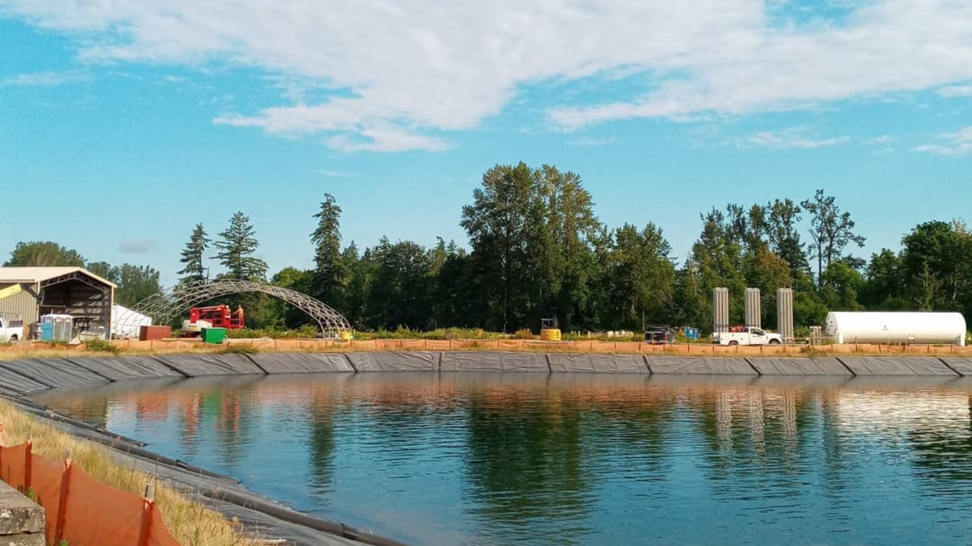 A wide, man-made pond with a black liner, surrounded by dirt banks, industrial structures, and a treeline under blue sky.