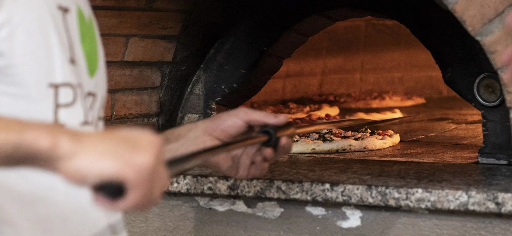 Una persona che mette le pizze in un forno di mattoni con un lungo attrezzo; dall'interno emana un caldo bagliore.