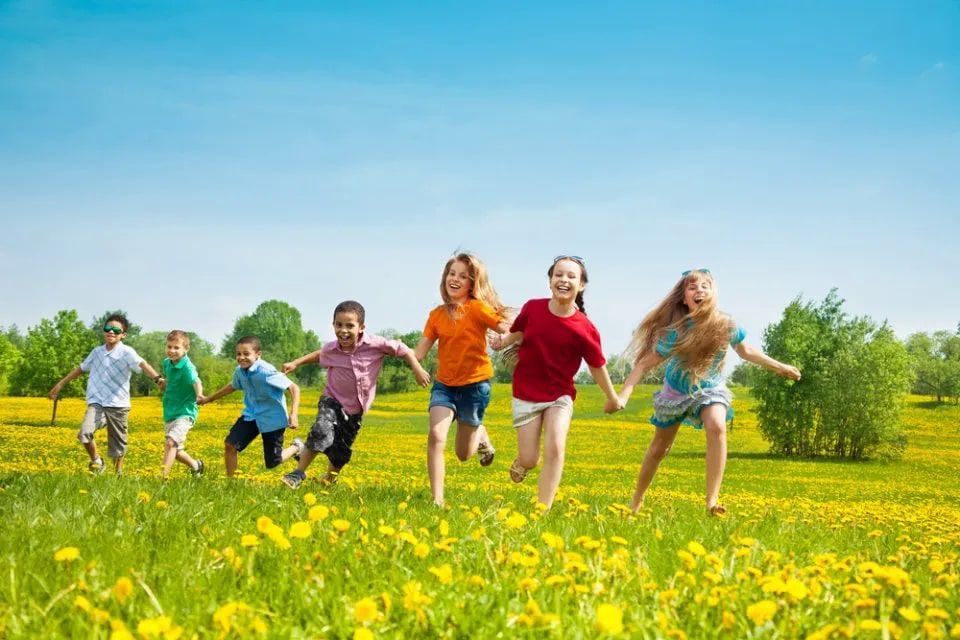 Bambini che corrono e ridono in un campo soleggiato e fiorito sotto un cielo azzurro.