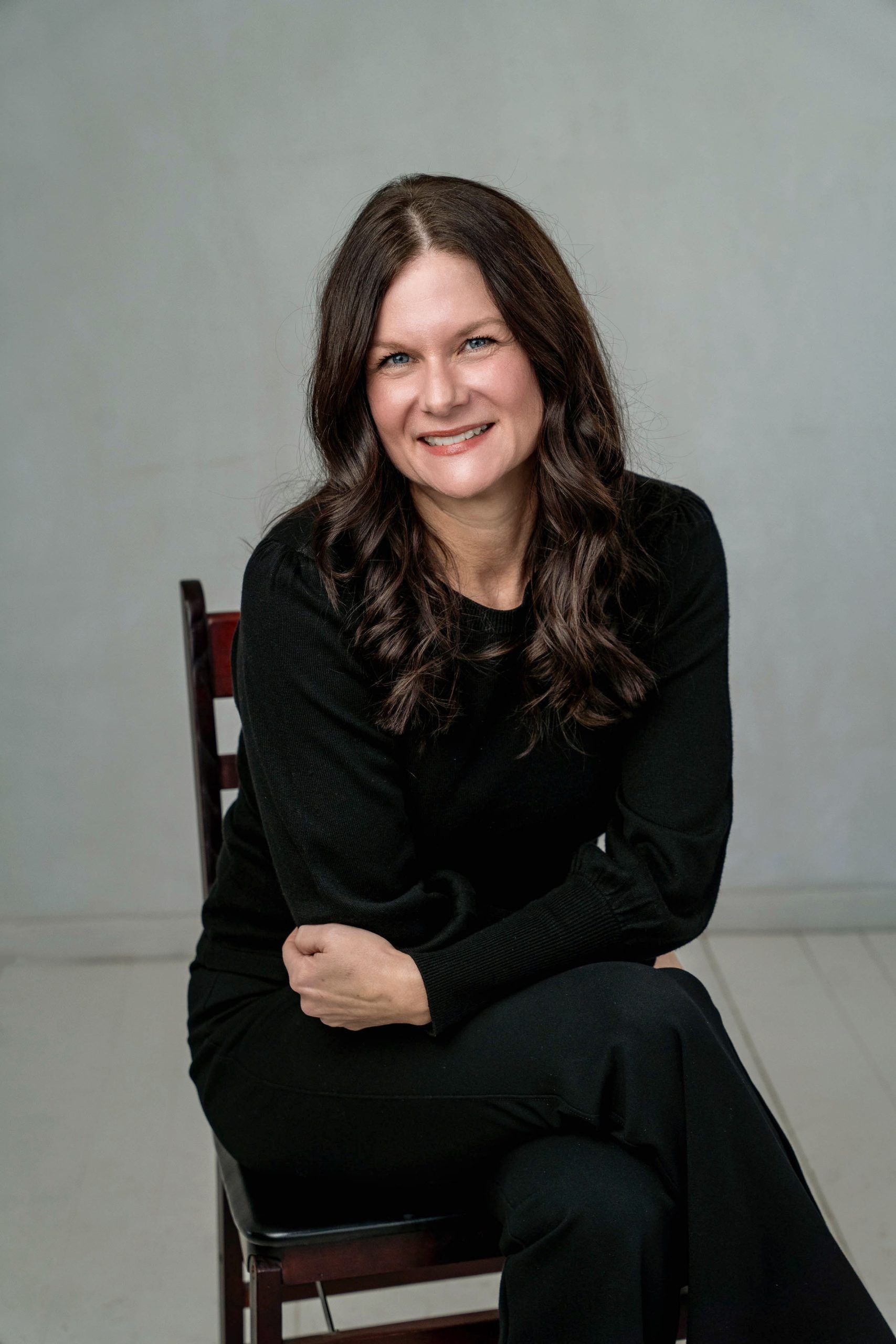 Smiling person in black outfit seated on a chair against a gray backdrop