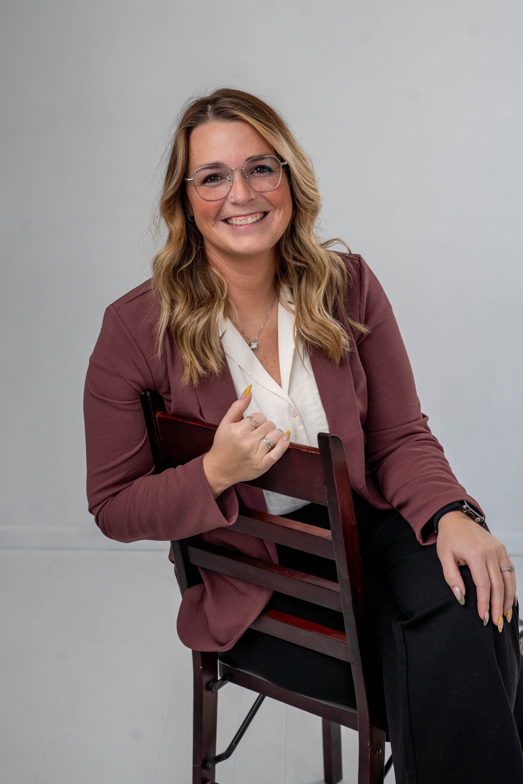 Woman seated on a chair, smiling and holding a notebook against a plain gray background.